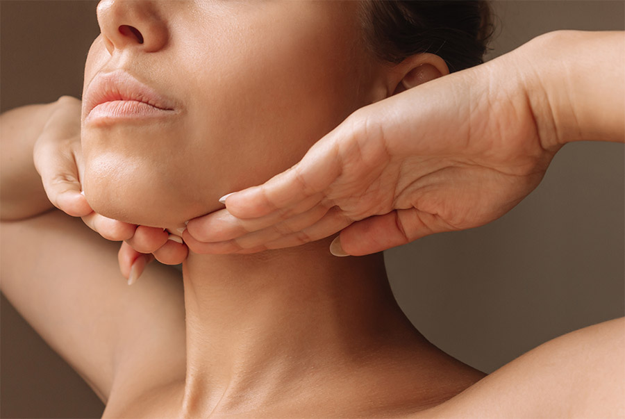 Cropped shot of young caucasian woman touching under the chin with hands massaging her face on dark brown background. Rejuvenation, facelift, facefitness. Exercises from the second chin, pelican neck